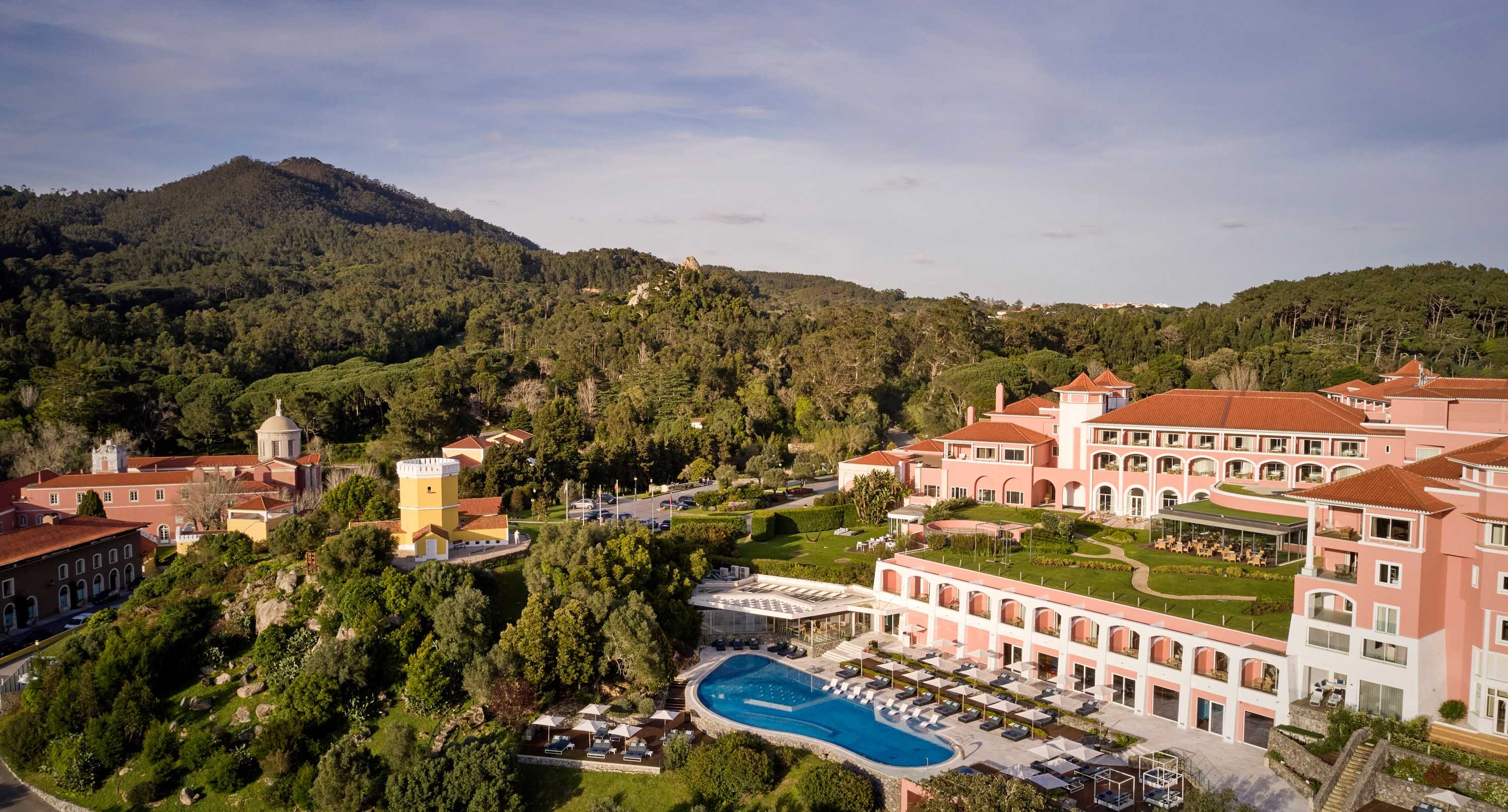 Aerial view of Penha Longa Resort at golden hour: the pink hotel, pool and monastery below the wooded Sintra hills